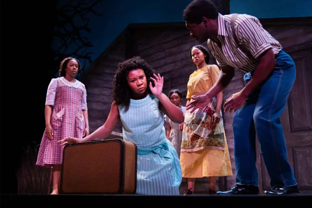 Four people stand around a woman kneeling beside a suitcase on stage; the scene suggests a dramatic moment in a theatrical performance.