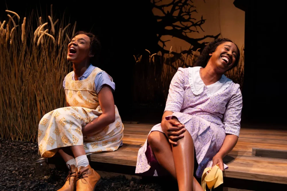 Two women in vintage dresses sit on a wooden porch, smiling and laughing, with wheat stalks and a silhouetted tree in the background, under stage lighting.