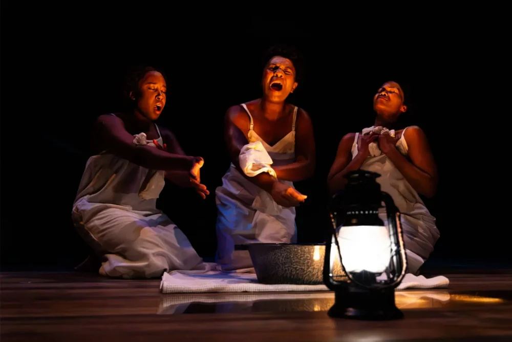 Three women in white dresses kneel onstage, dramatically gesturing around a metal basin, illuminated by a lit lantern in the foreground.