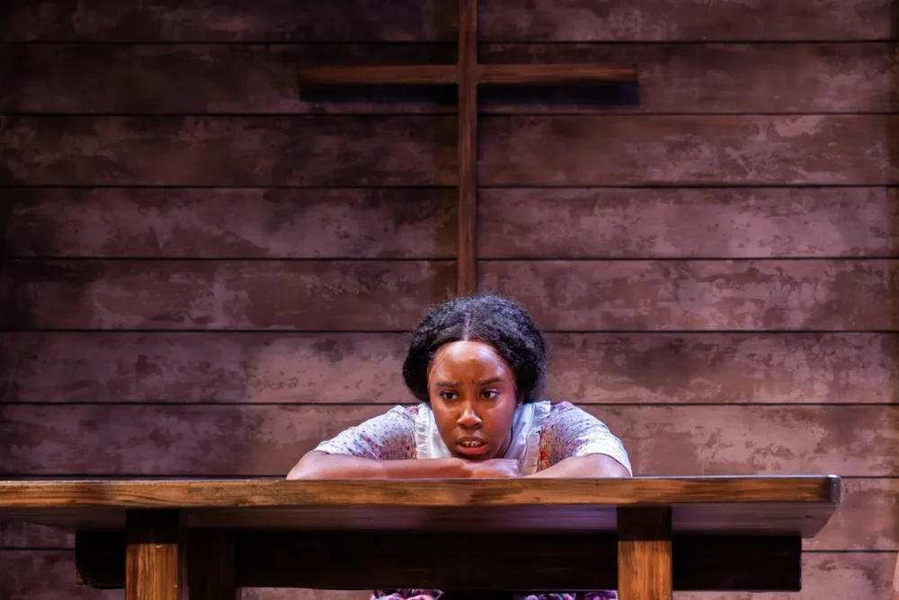 A woman leans on a wooden table, looking concerned, with a wooden cross on the wall behind her in a dimly lit room.