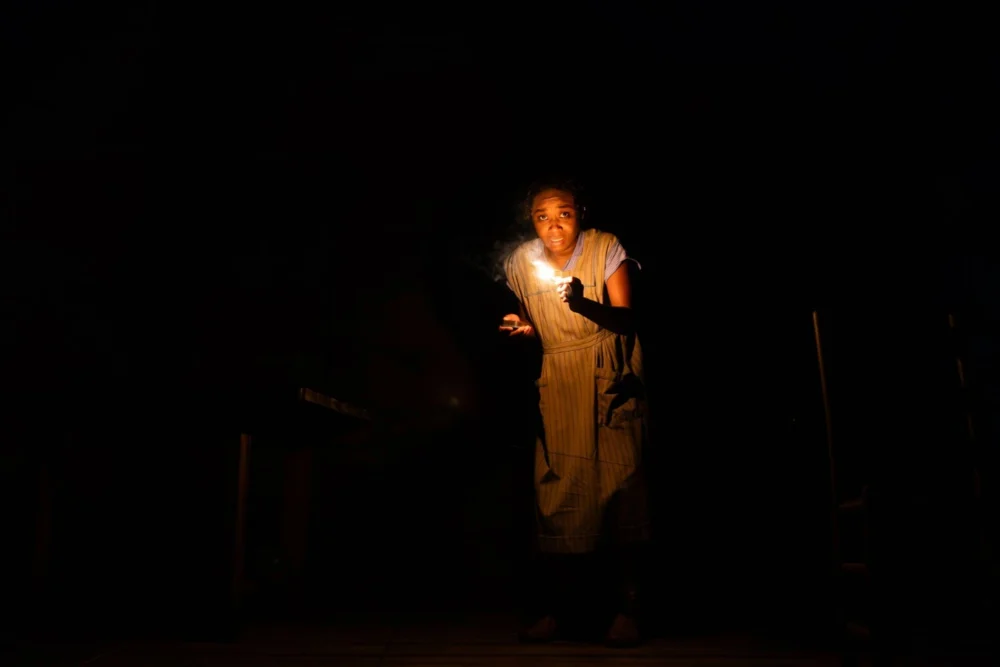 A person in a striped dress stands in darkness, holding a lit candle that illuminates their face and upper body.