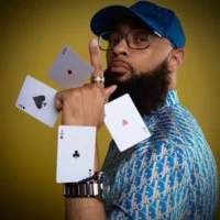 A man wearing glasses and a blue patterned shirt poses with four playing cards floating in the air around his raised hand against a yellow background.