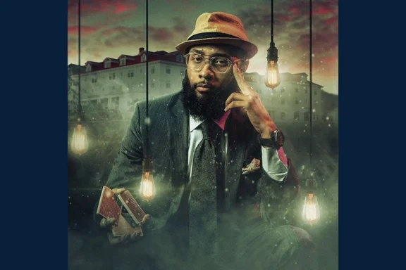 A man in a suit and hat sits holding a deck of cards, surrounded by hanging light bulbs, with a large building in the background and mist at his feet.