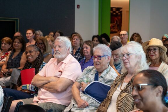 A diverse group of adults sits attentively in a well-lit room, listening to a speaker or presentation.