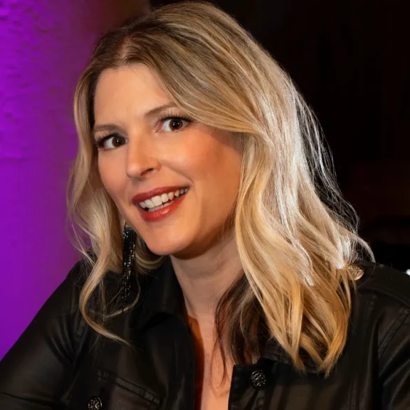 A woman with long, wavy blonde hair and a black leather jacket smiles at the camera against a purple-lit background.