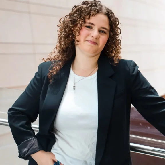 A person with curly hair wearing a black blazer and white shirt stands indoors, leaning on a railing and looking at the camera.