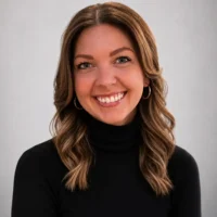 A woman with wavy brown hair, wearing a black turtleneck and hoop earrings, smiles at the camera against a plain light gray background.