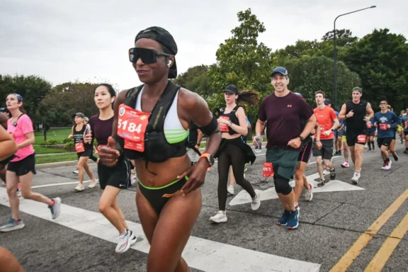 A group of marathon runners move along a city street, each wearing athletic clothing and numbered race bibs, with trees and greenery visible in the background.