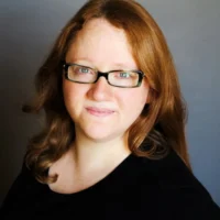 A woman with long reddish-brown hair and glasses is wearing a black top and smiling slightly at the camera against a plain background.