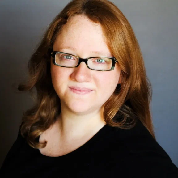 A woman with long reddish-brown hair and glasses is wearing a black top and smiling slightly at the camera against a plain background.