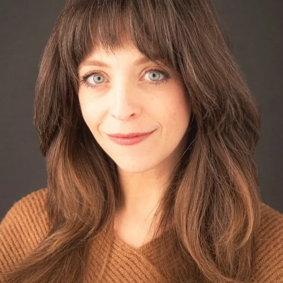 Woman with long brown hair and bangs, wearing a brown textured top, smiling softly against a plain dark background.