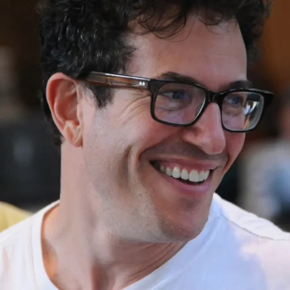 A man with curly dark hair and glasses smiles while looking to the side, wearing a white shirt.