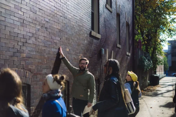 A man gestures toward a tall brick wall while speaking to a small group of people standing in an alleyway on a sunny day.