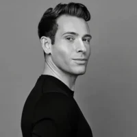 A man with short, styled hair wearing a black shirt stands against a plain gray background, looking over his shoulder at the camera.