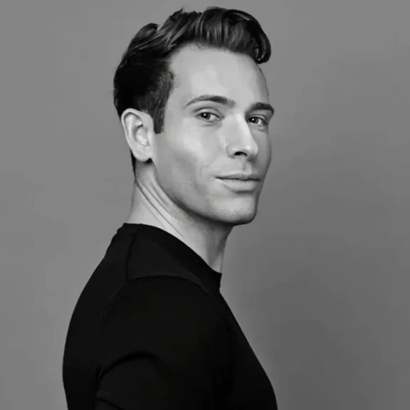 A man with short, styled hair wearing a black shirt stands against a plain gray background, looking over his shoulder at the camera.