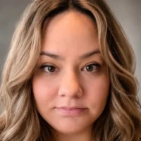 A woman with wavy, light brown hair and a neutral expression looks directly at the camera against a plain background.