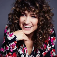 A woman with curly brown hair wearing a colorful floral-patterned blouse smiles while resting her chin on her hand against a dark background.