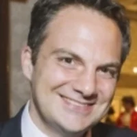 A man in a suit smiling at the camera in an indoor setting with blurred people and lights in the background.