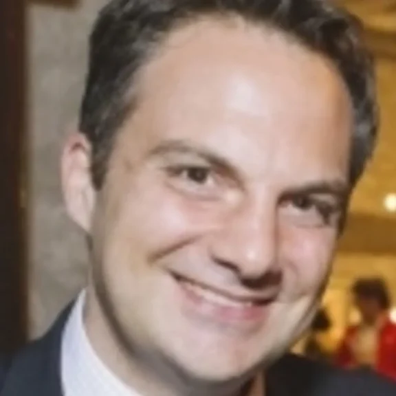 A man in a suit smiling at the camera in an indoor setting with blurred people and lights in the background.