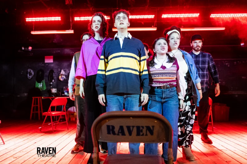 A group of six actors stands closely together on a stage with red lighting, facing forward. Empty chairs, including one labeled "Raven," are visible in the foreground.