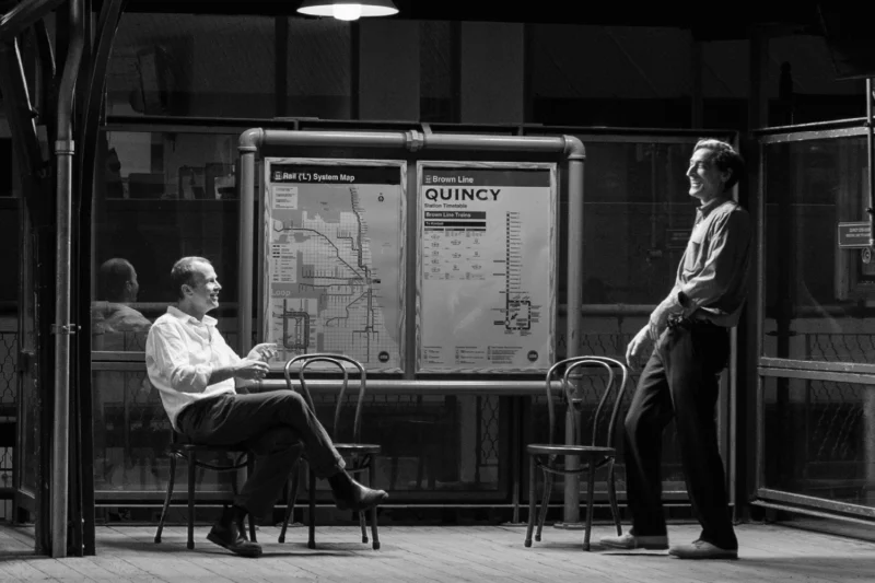 Two men converse on a train platform at night, with chairs and transit maps labeled "Quincy" visible behind them. The scene is lit by overhead lights.