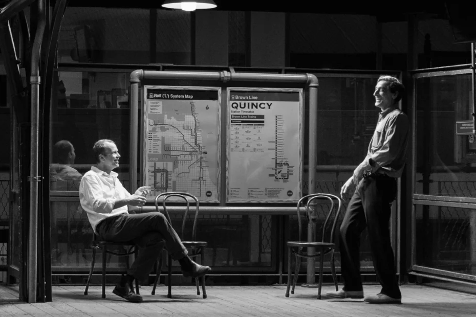 Two men converse on a train platform at night, with chairs and transit maps labeled "Quincy" visible behind them. The scene is lit by overhead lights.