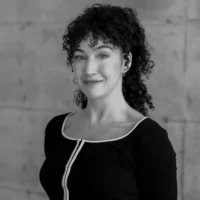 A person with curly hair wearing a black top with a white trim stands in front of a plain concrete wall, looking at the camera and smiling slightly.