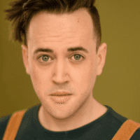 A person with short, styled brown hair and green eyes looks directly at the camera, wearing a green shirt and brown suspenders against a plain green background.