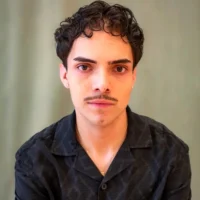 A young person with short curly hair, a mustache, and earrings wears a black collared shirt, facing the camera against a plain, light-colored background.