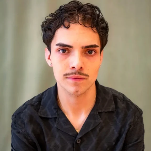 A young person with short curly hair, a mustache, and earrings wears a black collared shirt, facing the camera against a plain, light-colored background.