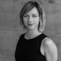 A woman with short, light hair wearing a sleeveless dark top and a necklace stands in front of a plain, blurred background, looking at the camera.