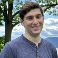 A person with short brown hair wearing a blue henley shirt smiles outdoors, with trees, water, and a cloudy sky in the background.
