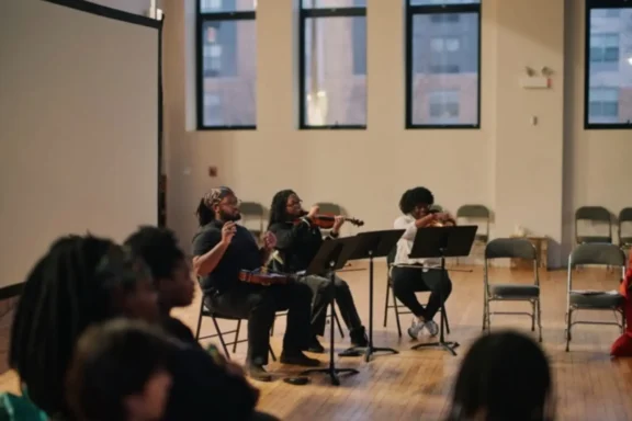 Three people sit and play string instruments in a well-lit room with large windows, while an audience watches. Empty chairs and music stands are visible.
