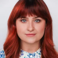 A woman with long, straight reddish-brown hair and blue eyes looks directly at the camera. She has a nose ring and wears a floral shirt. The background is plain white.