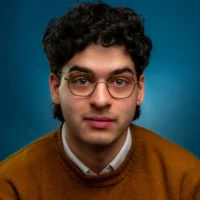 A young man with curly dark hair, glasses, and an earring, wearing a brown sweater over a white shirt, poses against a blue background.