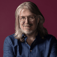 A middle-aged person with long gray hair, glasses, and a beard, wearing a blue checkered shirt, poses against a plain burgundy background.