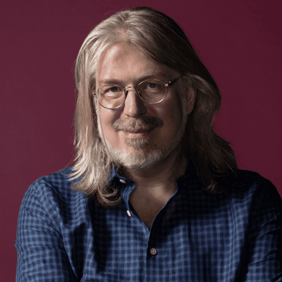 A middle-aged person with long gray hair, glasses, and a beard, wearing a blue checkered shirt, poses against a plain burgundy background.