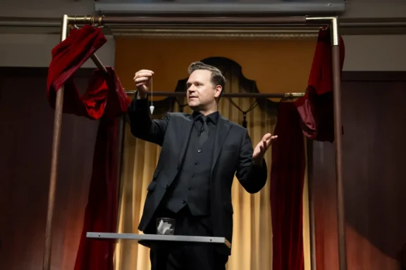 A man in a black suit performs a magic trick onstage, holding a small object in one hand, with red curtains and a table in front of him.