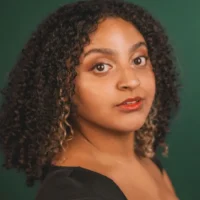A person with curly dark hair, light brown skin, and wearing a black top looks at the camera against a dark green background.