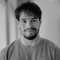 A man with wavy hair, a beard, and an earring is looking at the camera and smiling slightly. He is wearing a plain T-shirt. The image is in black and white.