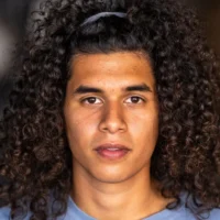 A person with long, curly hair pulled back by a headband, wearing a light blue shirt, looks directly at the camera with a neutral expression.