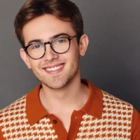A young adult with short brown hair, glasses, and a trimmed beard smiles at the camera, wearing an orange and cream patterned collared shirt against a gray background.
