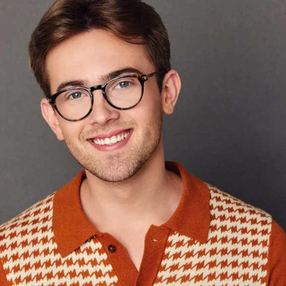 A young adult with short brown hair, glasses, and a trimmed beard smiles at the camera, wearing an orange and cream patterned collared shirt against a gray background.