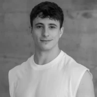 A young man with short dark hair wearing a sleeveless white shirt stands in front of a plain, textured background, looking at the camera and smiling slightly.