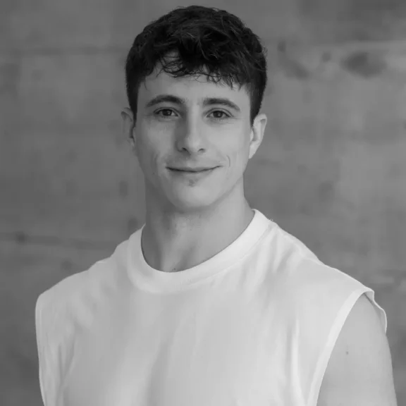 A young man with short dark hair wearing a sleeveless white shirt stands in front of a plain, textured background, looking at the camera and smiling slightly.