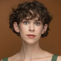 A person with short curly brown hair and light eyes looks directly at the camera against a plain brown background, wearing a green top.