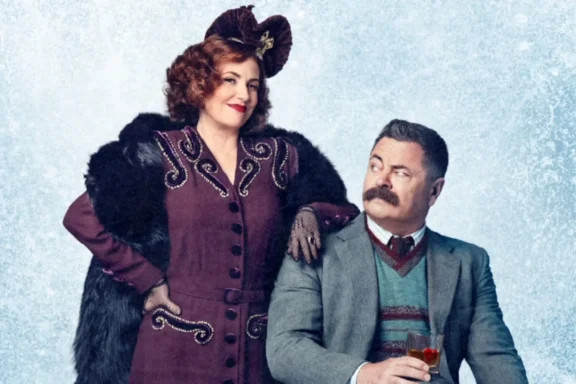 A woman in a vintage purple dress and hat stands beside a seated man in a suit holding a drink, both posing against a light blue background.