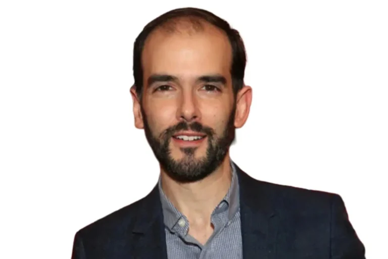 A man with a short beard and receding hairline is wearing a blue blazer over a collared shirt, standing against a plain white background.
