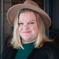 Person with shoulder-length blonde hair wearing a wide-brimmed tan hat, green top, and black jacket, standing indoors against a brick and metal background.