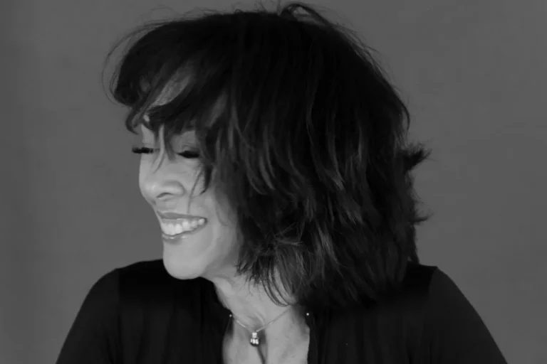 A woman with shoulder-length dark hair smiles with her eyes closed, facing left, against a plain background. She is wearing a dark top and a necklace.